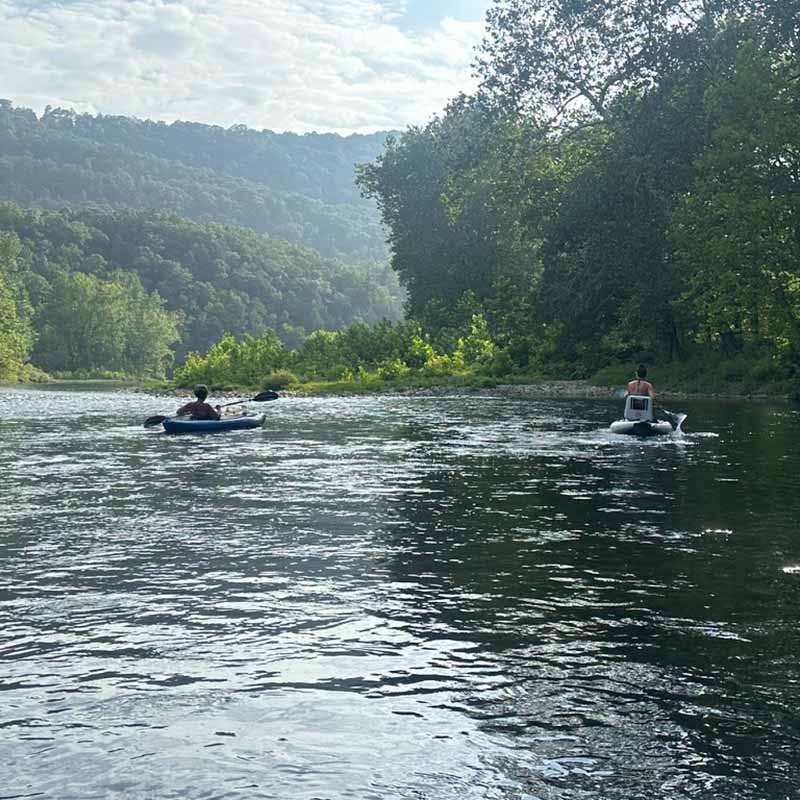 Kayakers paddling on the Cheat River in West Virginia, surrounded by forested hills under a partly cloudy sky.