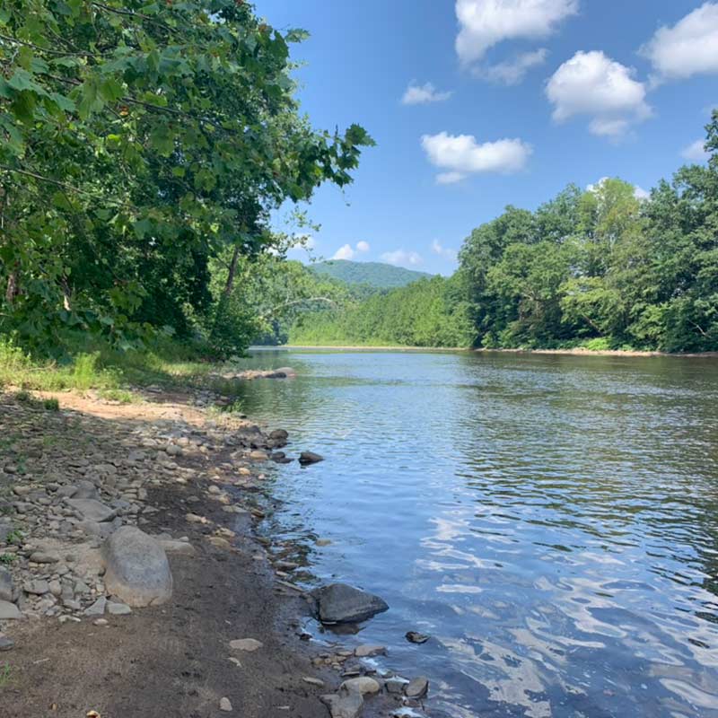 Cheat River riverfront lot in West Virginia with rocky riverbank, lush green trees, and blue sky reflected in calm water.